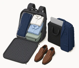An open charcoal travel backpack holding folded dress shirts and suit trousers in a dry-cleaner bag, with a navy suit jacket draped over a slim laptop bag beside it and brown slip-on dress shoes in front.