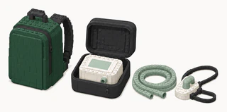 A green backpack beside an open black padded case holding a compact white breathing machine, with a coiled sage hose and a simple mask with black straps.